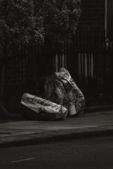 Two large black plastic rubbish bags, filled and tied at the top, are placed on the pavement beside a black metal fence with vertical bars. The bags appear to contain household waste or discarded mate