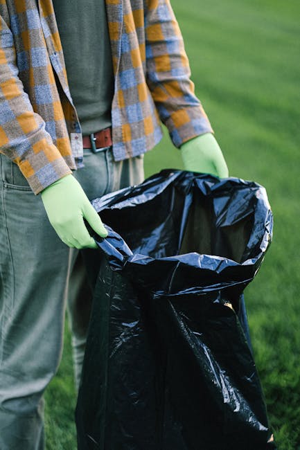 A person wearing a yellow, grey, and blue checked flannel shirt over a dark green T-shirt and grey trousers, standing outdoors on a grassy area. They are holding open a black plastic rubbish bag with 