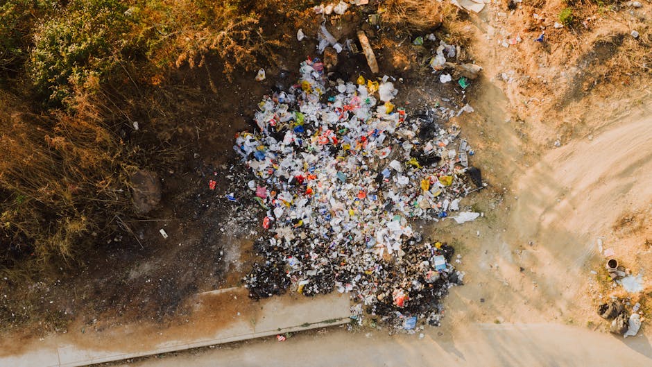 A black, cylindrical public waste bin with a domed lid, situated on a cobblestone street, overflowing with litter including empty beer bottles, crumpled paper, and food containers. Several discarded b